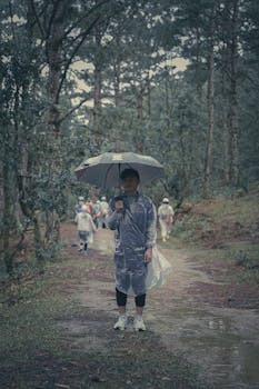 A child wearing a raincoat holds an umbrella standing on a forest path. Rainy day hike.