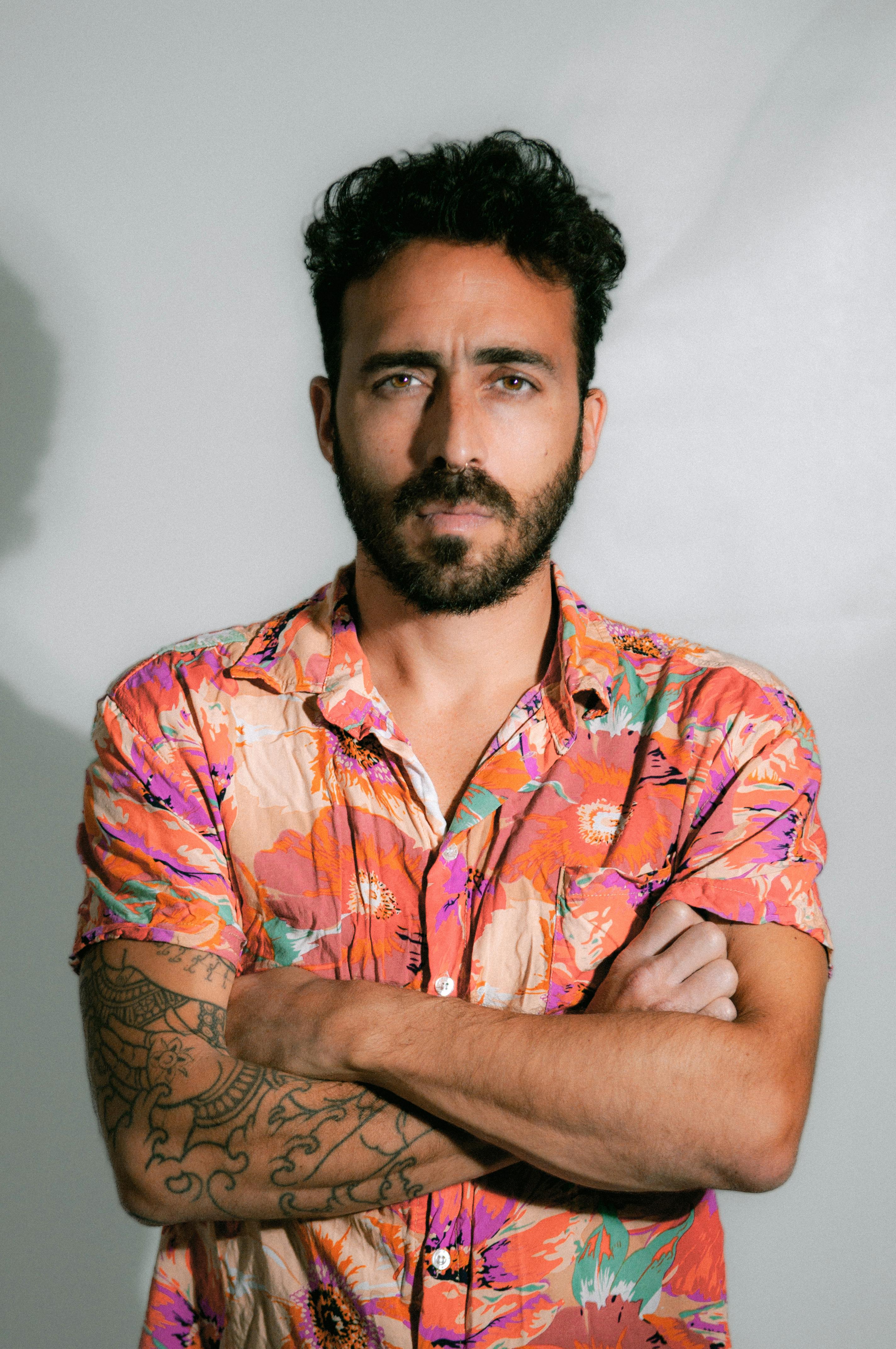 Man with a colorful shirt and tattoos posing confidently in a studio setting.