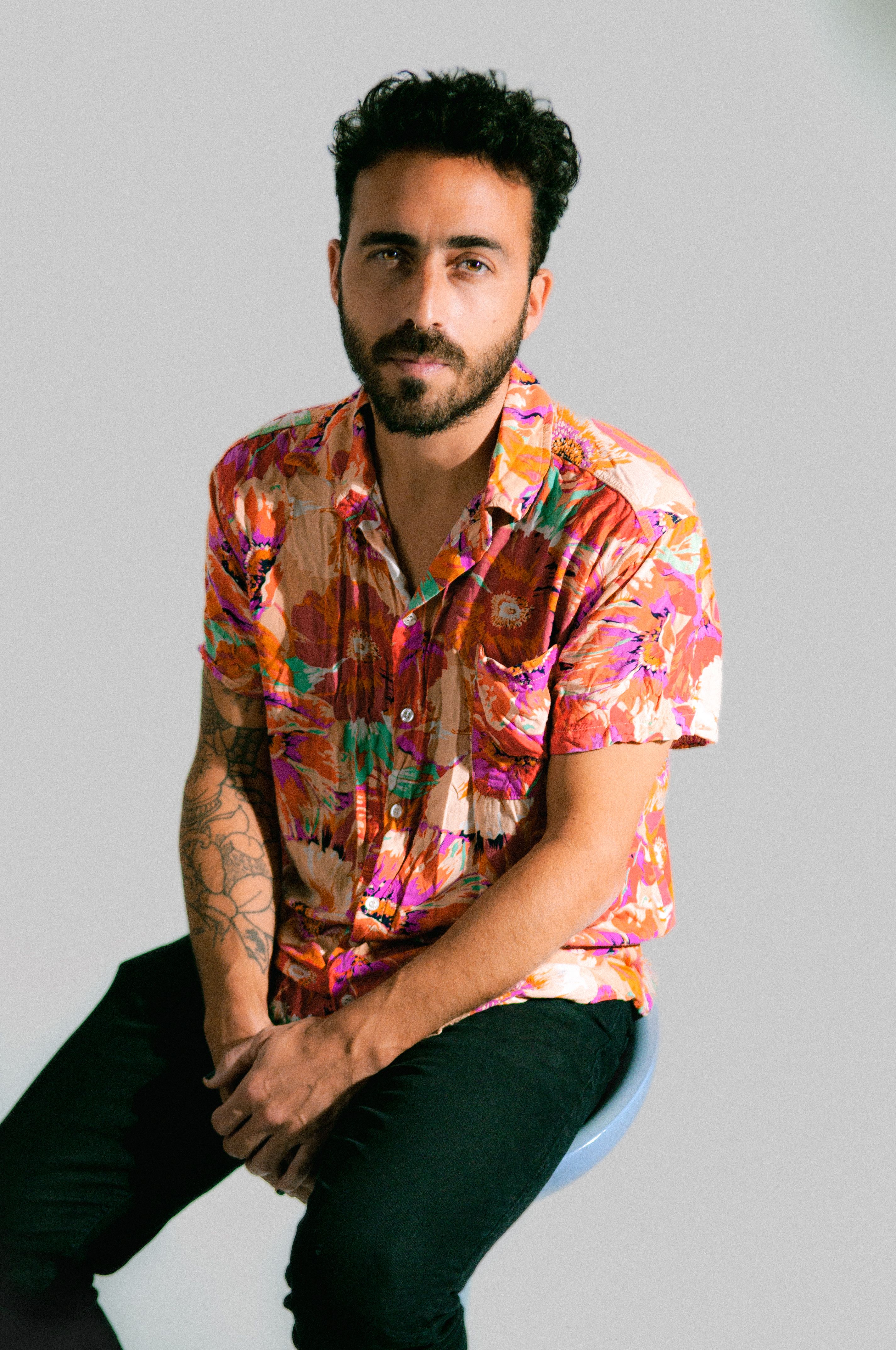 Portrait of a man in a vibrant, floral shirt sitting in a studio setting, highlighting fashion and tattoos.