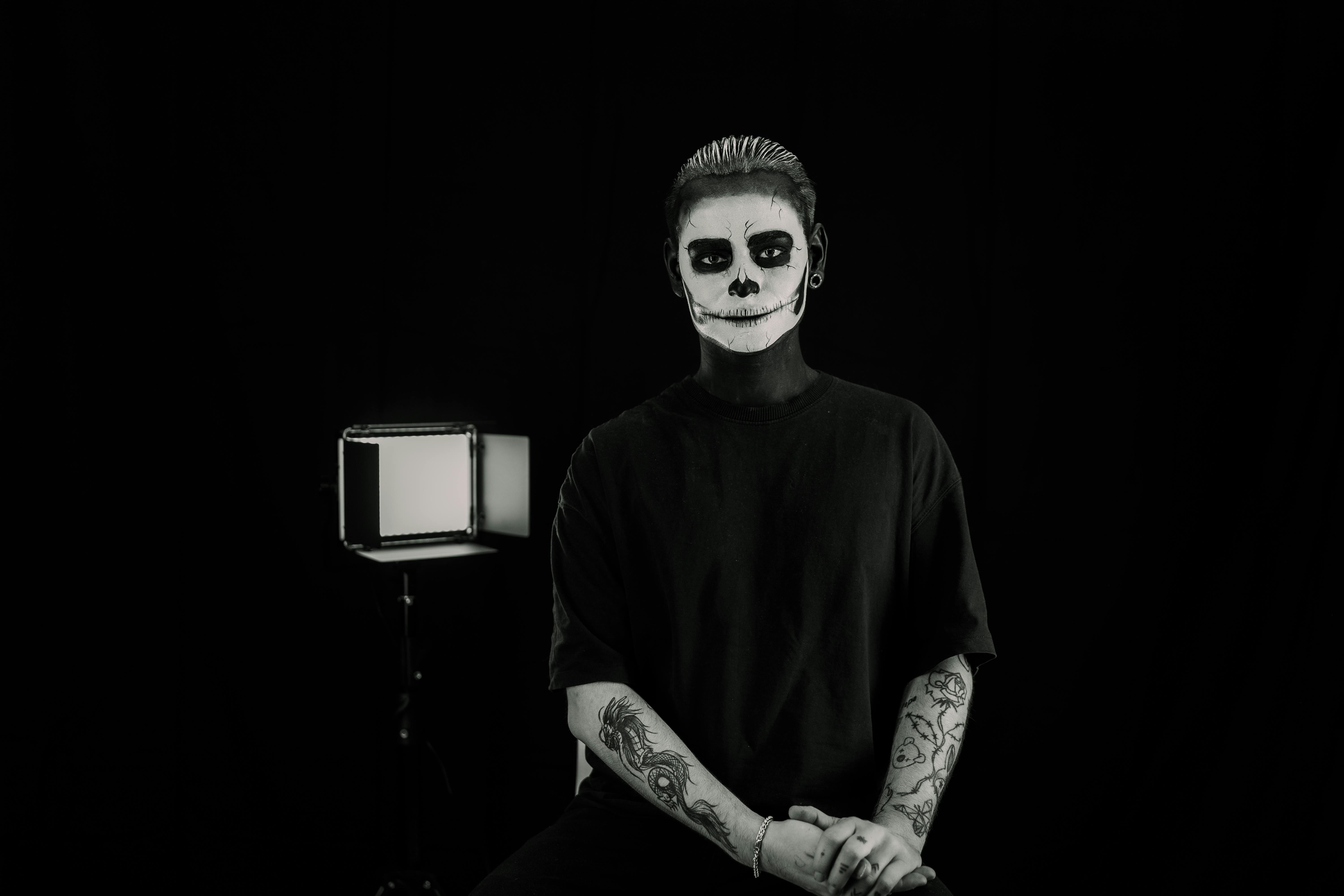 A black and white portrait showcasing a person with skull face paint and tattoos in an ominous setting.