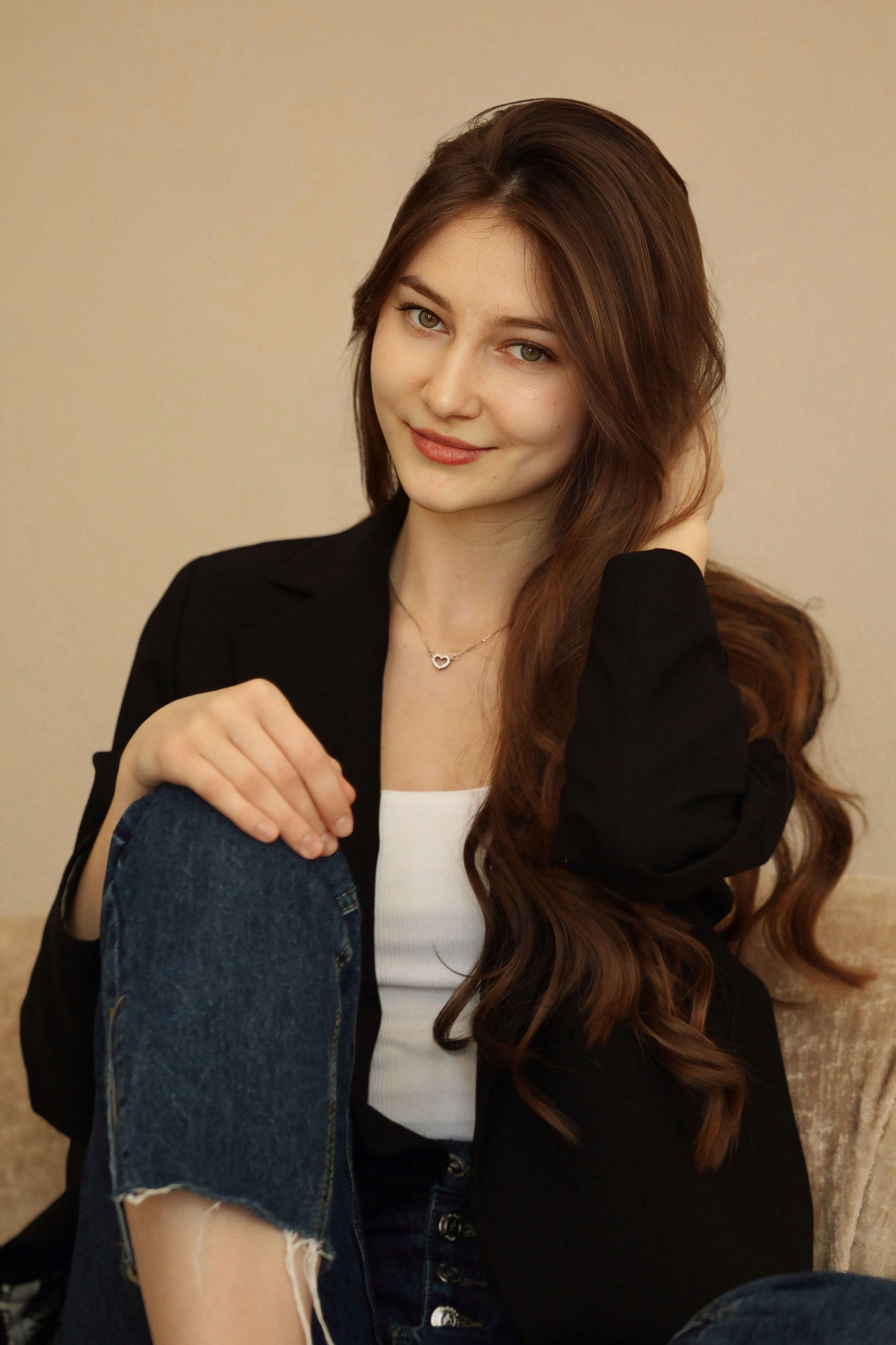Stylish woman with long brown hair sitting indoors on a beige couch, smiling elegantly.