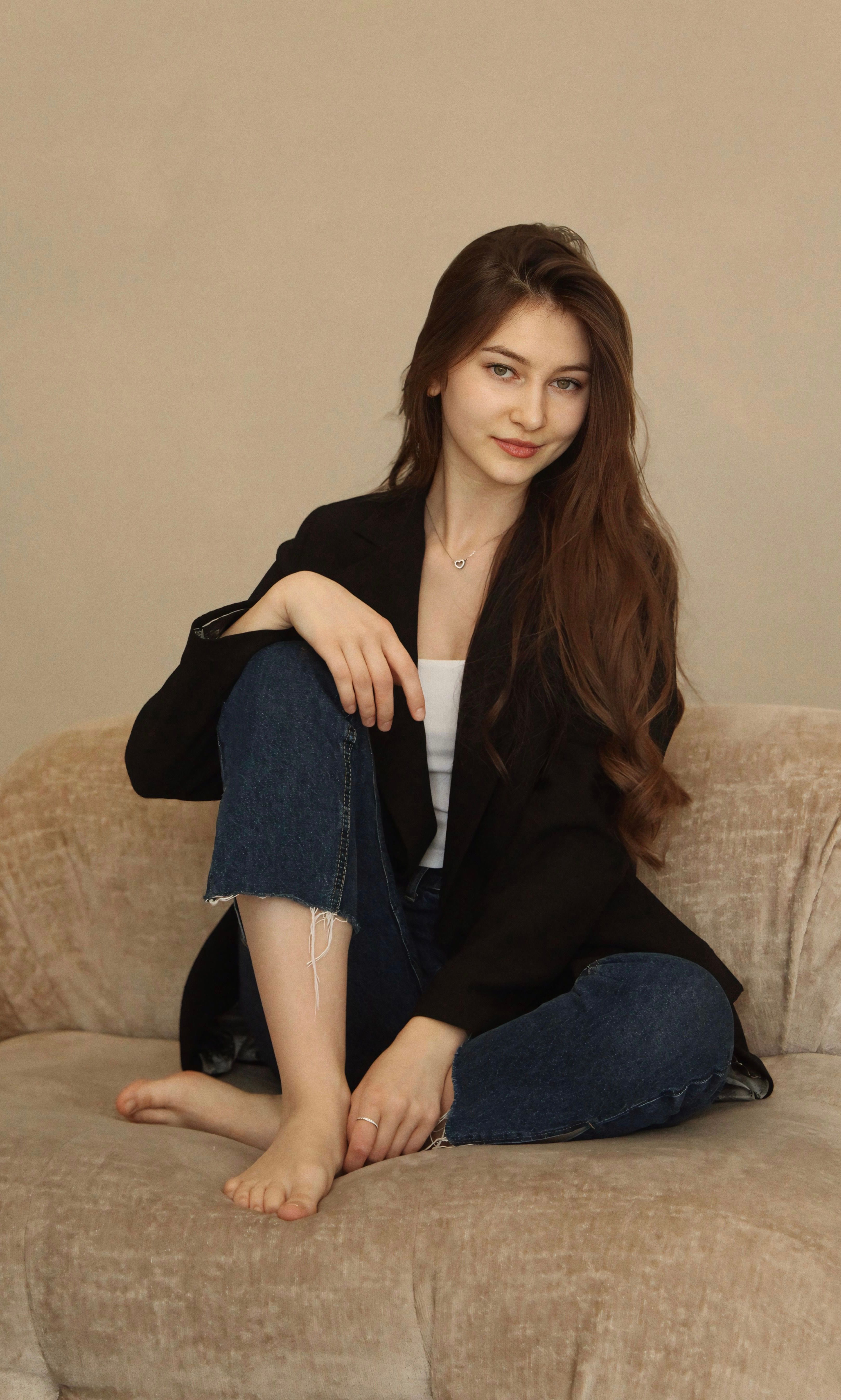 Elegant woman in casual outfit sitting barefoot on a couch, smiling gently.