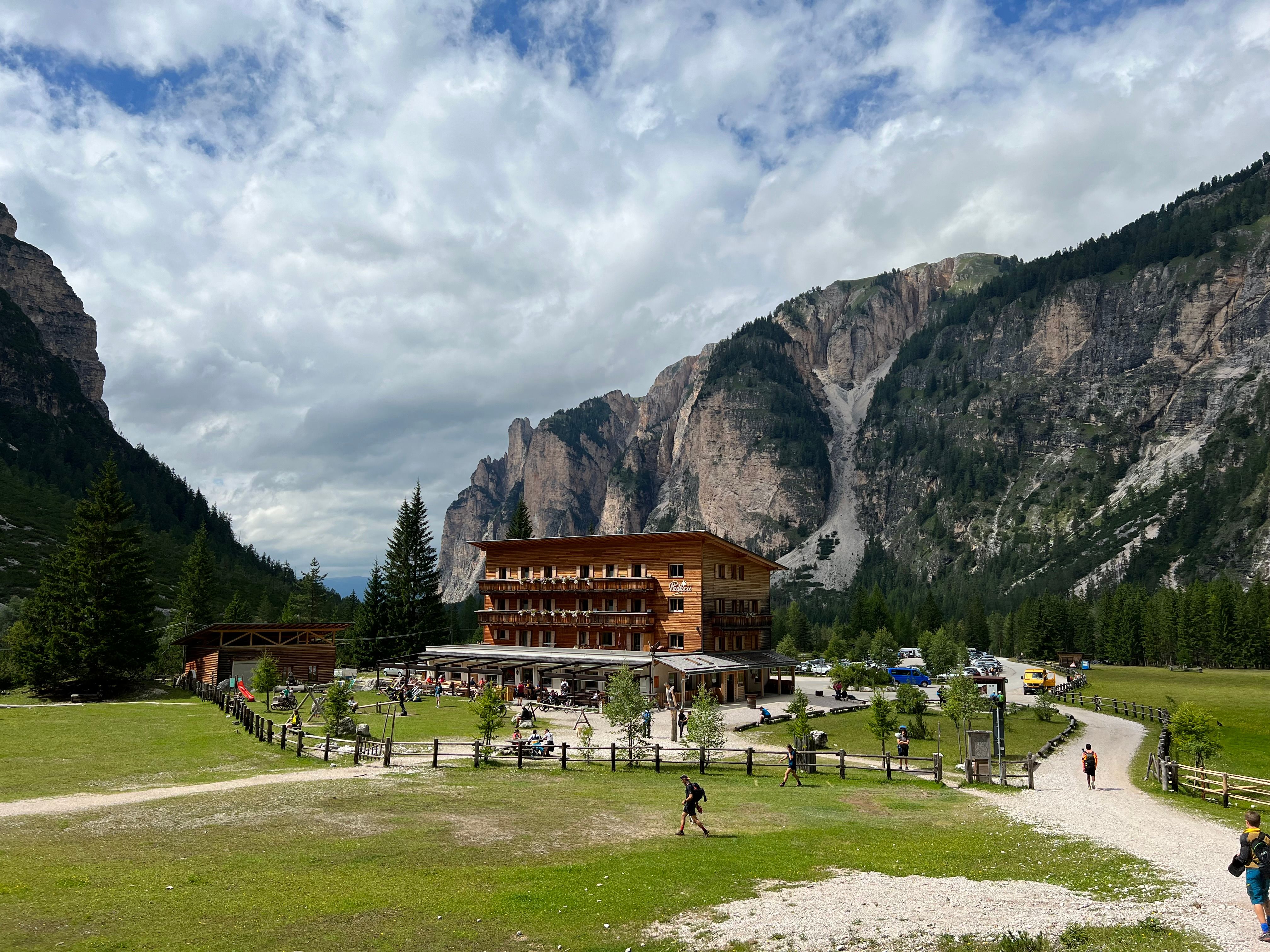A rustic wooden lodge nestled in a lush alpine valley, surrounded by majestic mountains and scattered clouds.