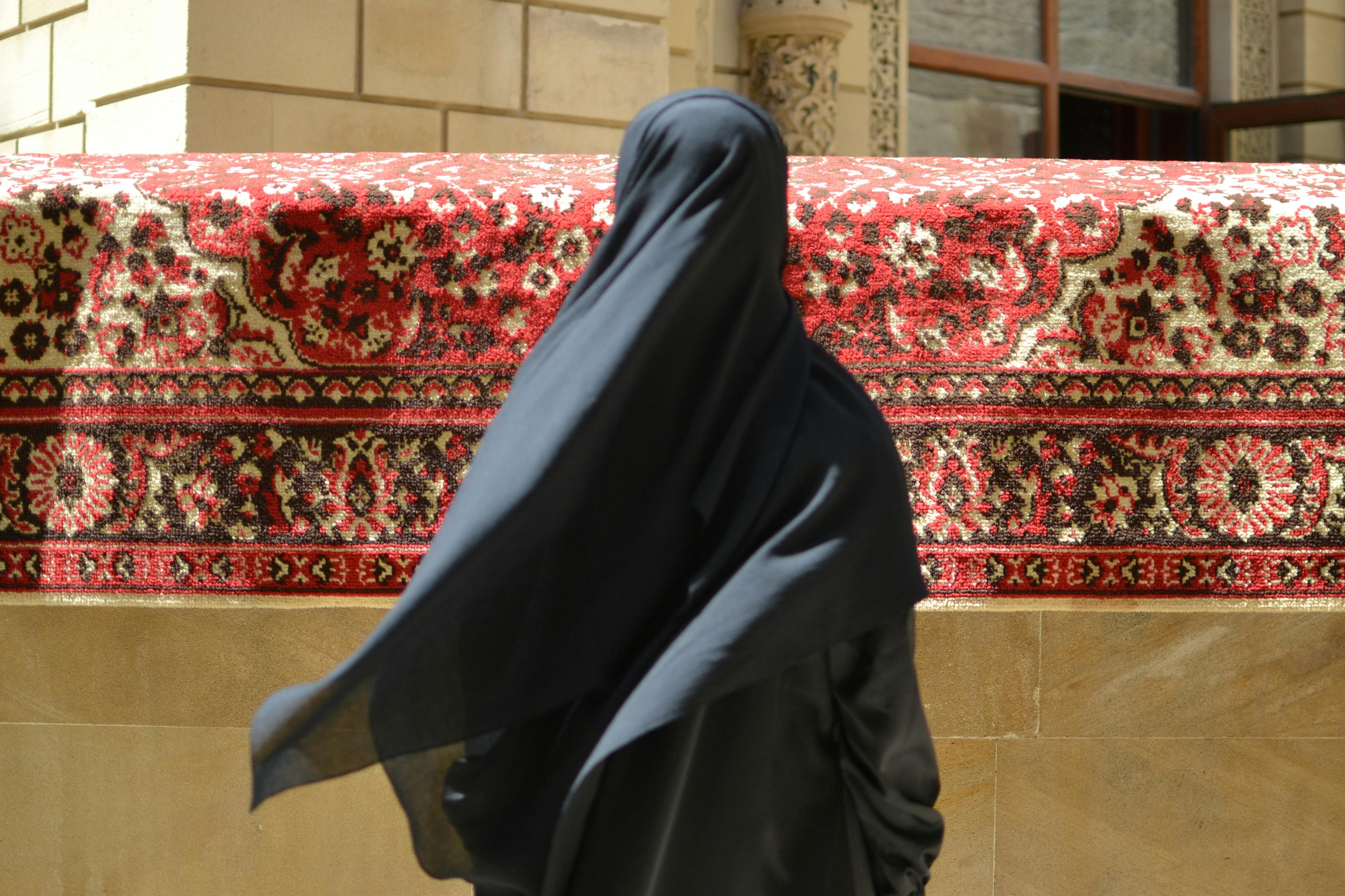 abaya next day delivery - A woman in a black abaya walking past a traditional Persian carpet in an urban setting.