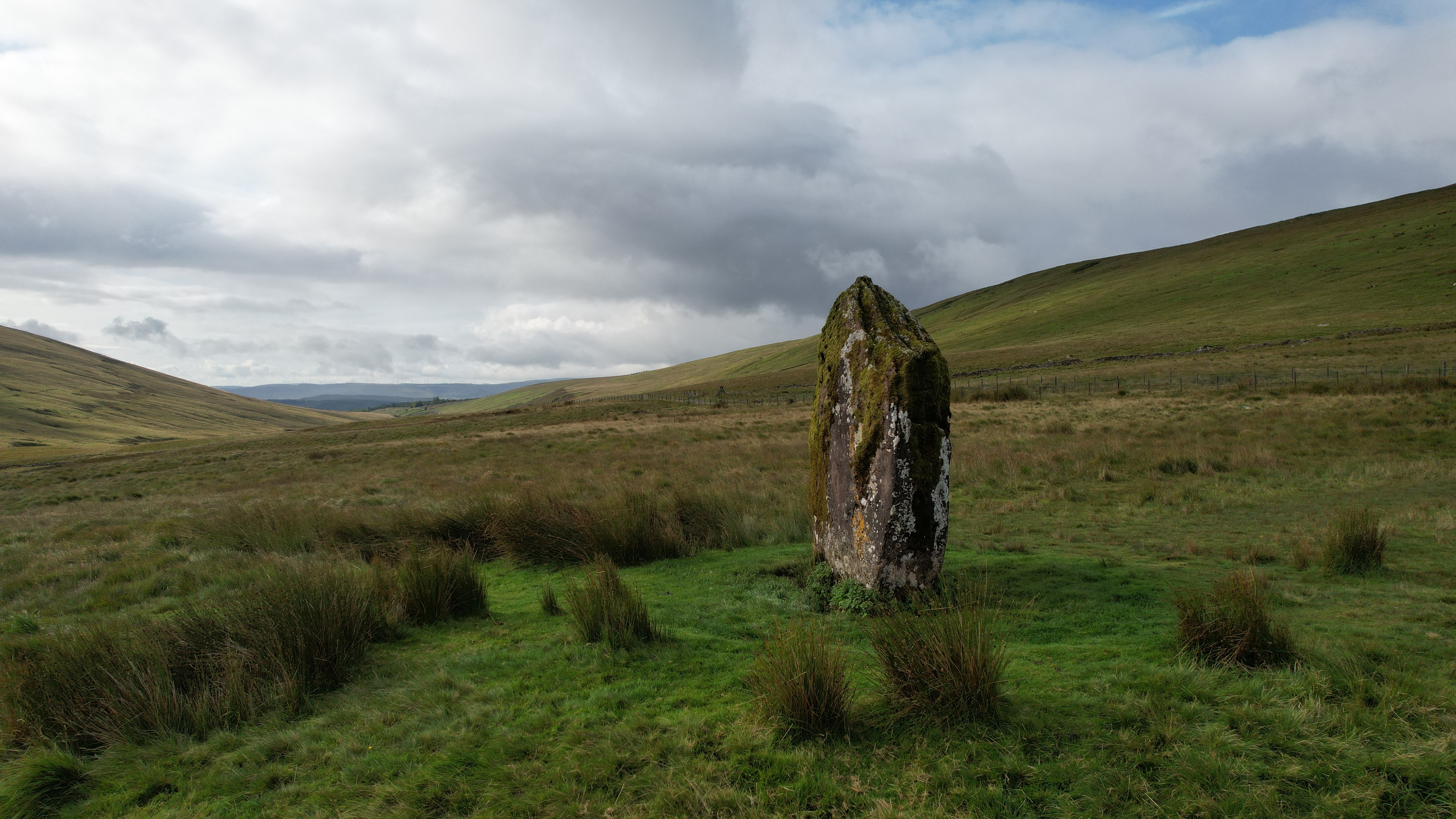 A solitary stone monolith stands in the green grasslands of Usk, Wales under a cloudy sky.