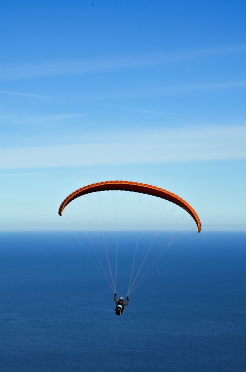 Free A person paragliding with an orange chute over a vast ocean under clear blue skies. Stock Photo