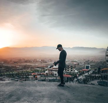 A man in a cap stands on a rooftop, overlooking a scenic cityscape during a beautiful sunset.