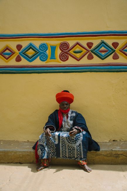 A man in traditional clothing sits against a colorful decorative wall, showcasing cultural attire.