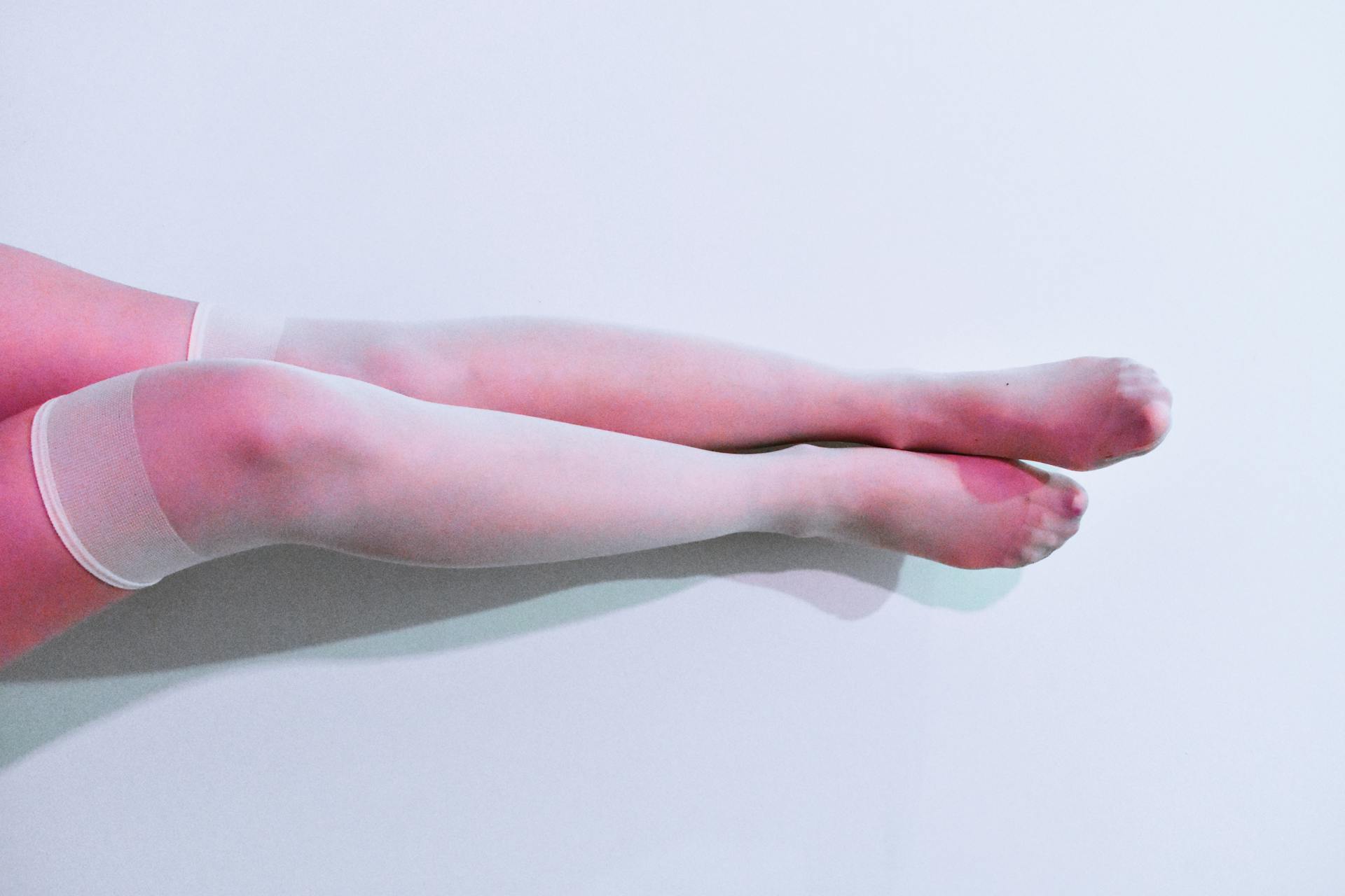 Close-up of female legs in white stockings on a pastel pink backdrop, creating a modern and minimalist aesthetic.