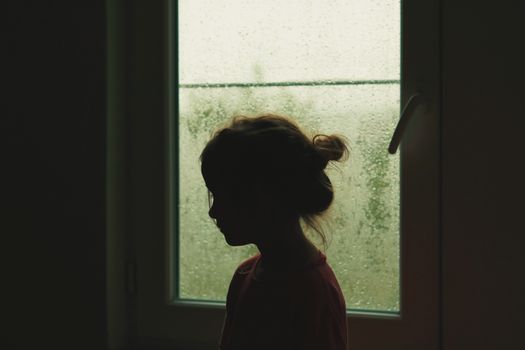 A child's silhouette stands by a rainy window, creating a nostalgic indoor scene.