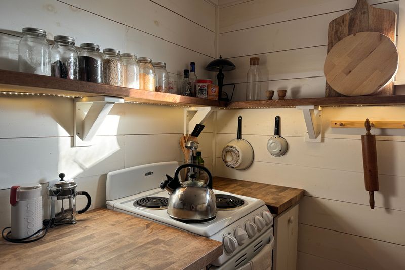 A small rustic kitchen with a wooden shelf, stove, and cooking utensils, offering a warm and cozy atmosphere.