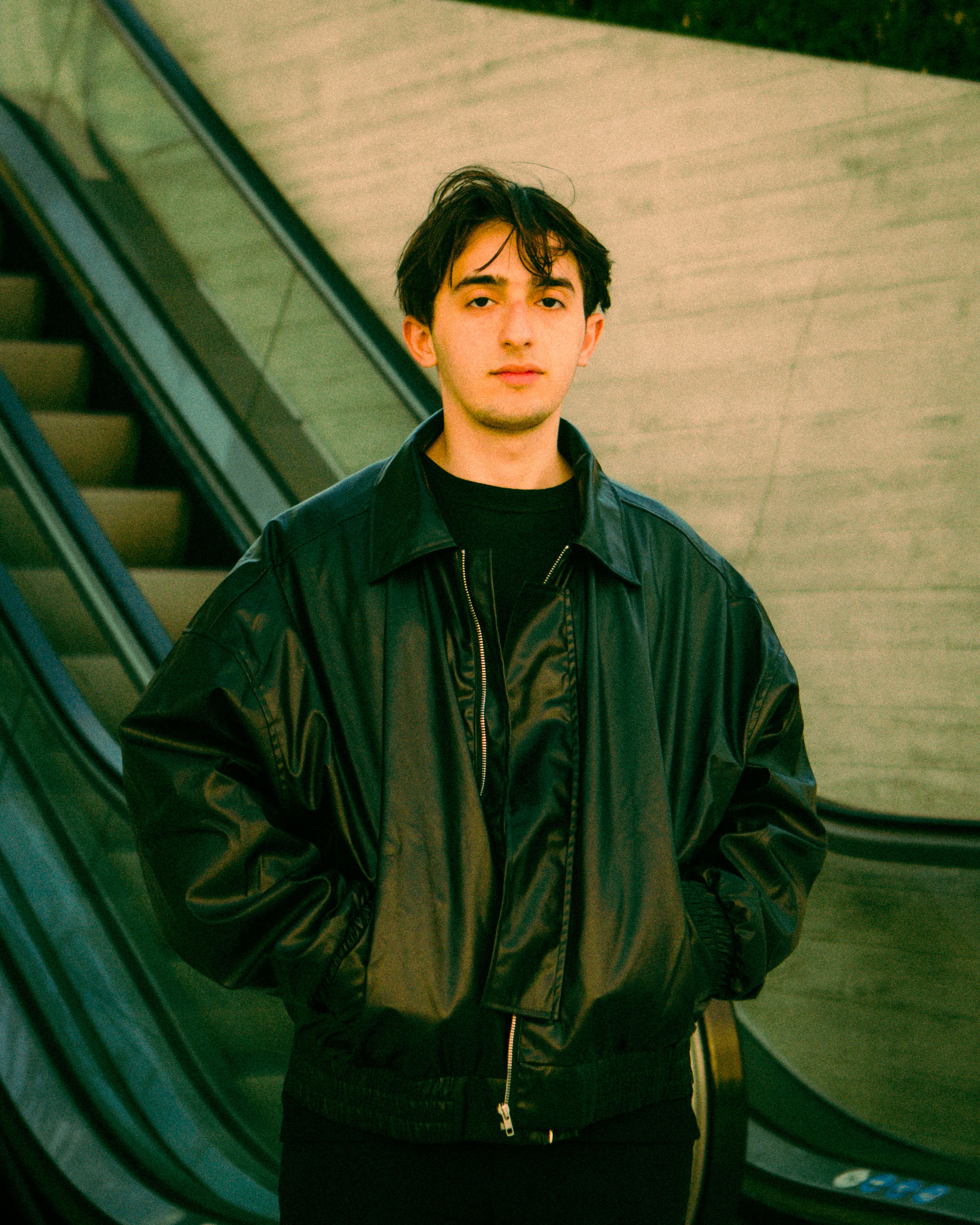 Portrait of a young man in a leather jacket standing by escalators in an urban environment.
