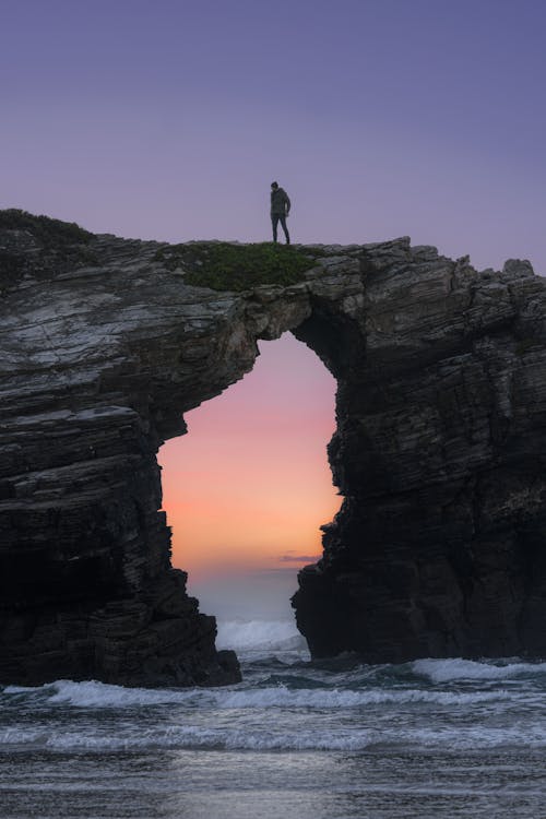 Man Standing on Natural Arch over Sea Shore at Dusk