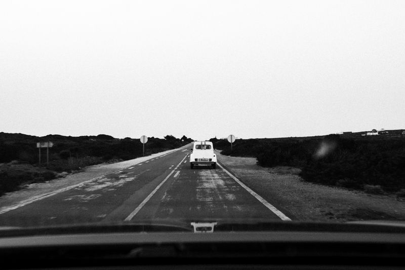 A black and white photo of a vintage car driving on an empty rural highway.