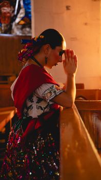 A woman in colorful Mexican dress prays in a sunlit church in Huauchinango, Mexico.