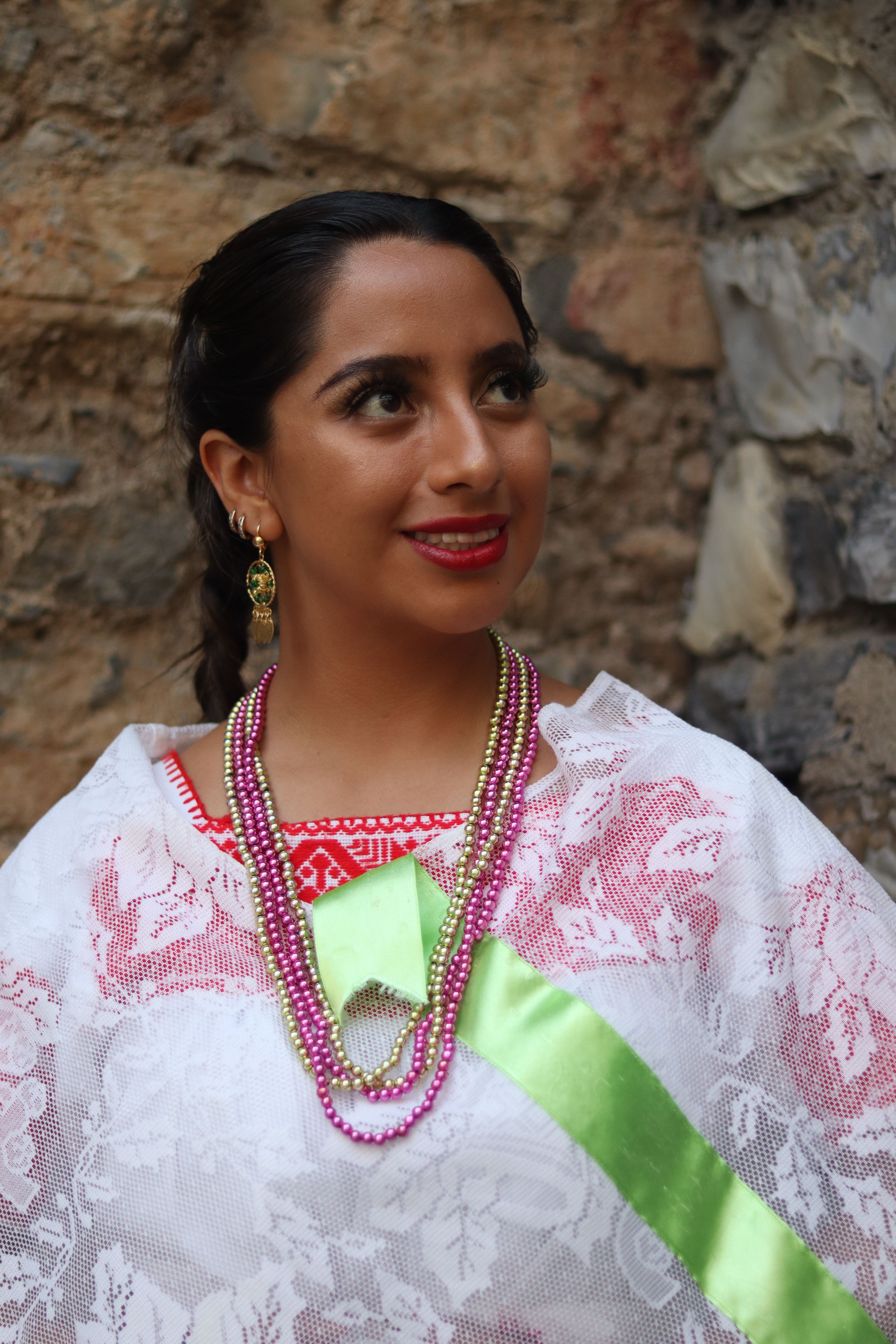 A woman in traditional dress with lace and colorful jewelry, posing outdoors against a stone wall.