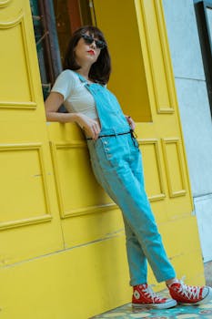 一位戴着墨镜、身穿工作服的时尚女士，倚在充满活力的黄色墙壁上，展现出休闲的城市风格