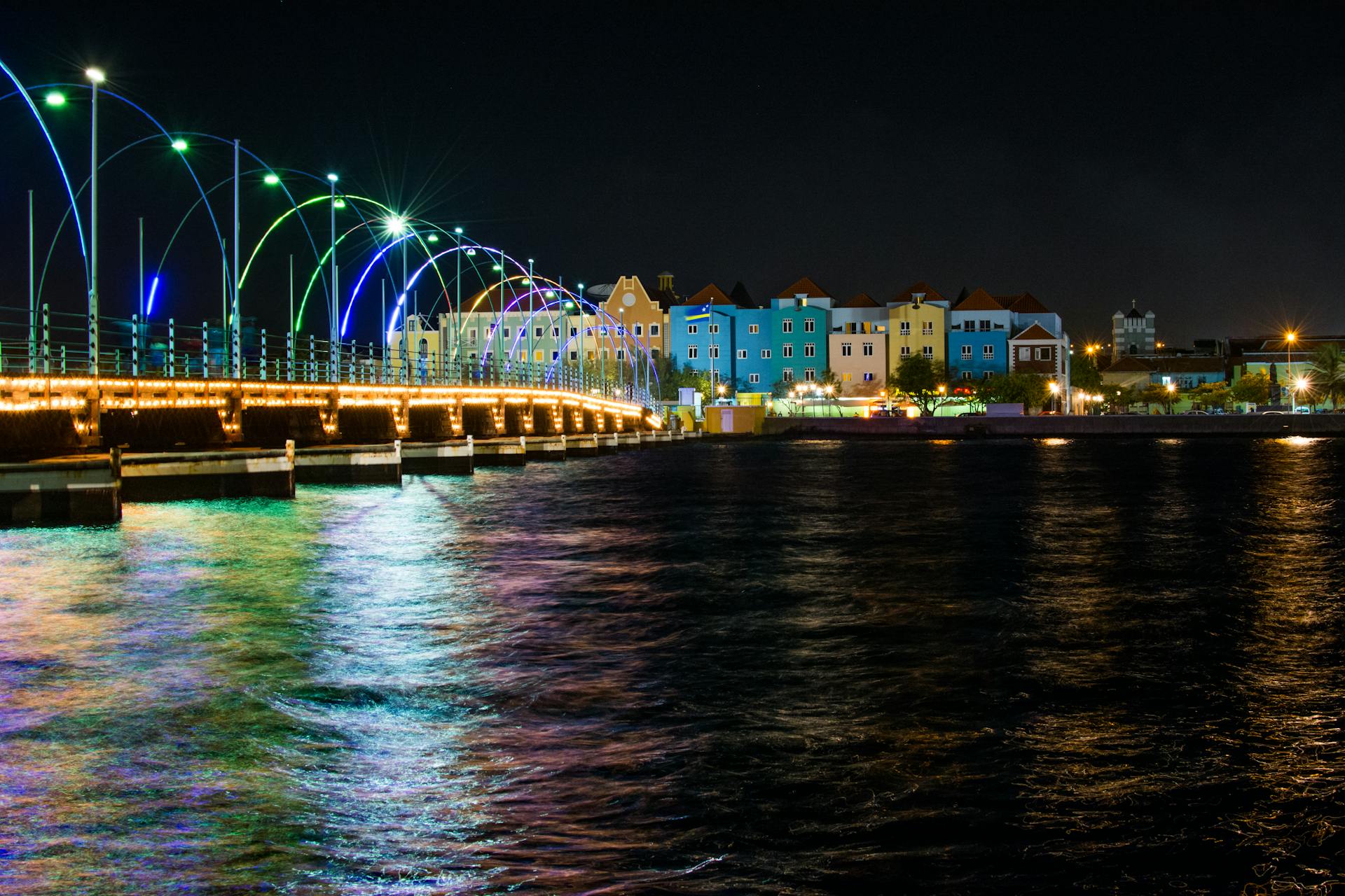 Vibrant night view of the Queen Emma Pontoon Bridge in Curacao with colorful lights and waterfront reflection.