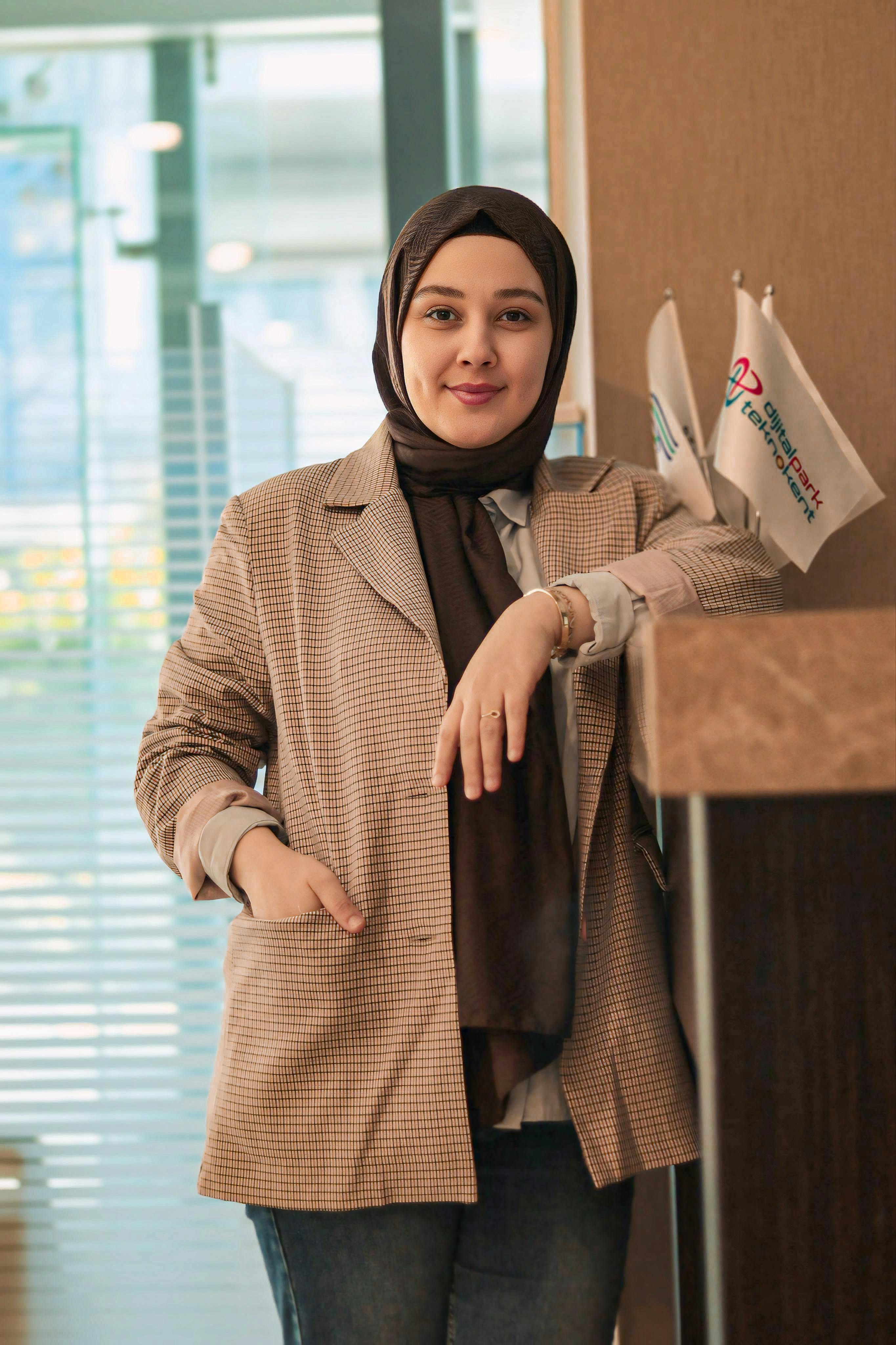 A confident woman standing in an office, wearing a hijab and checkered blazer, exuding professionalism.