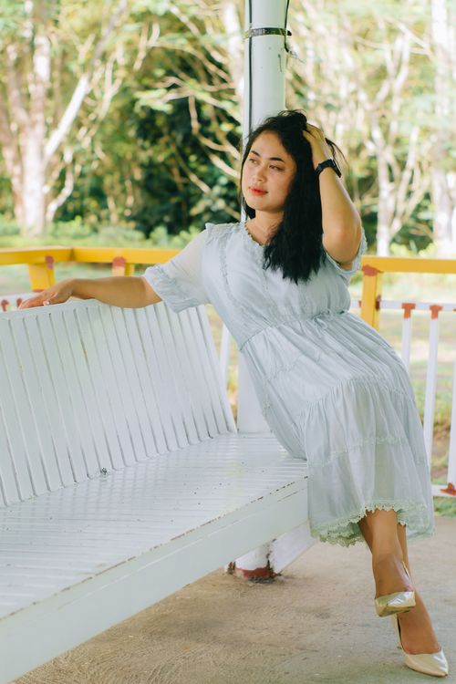 Free A woman in a white sundress sits on a bench outdoors, enjoying a sunny day. Stock Photo