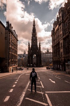 A street view of the Scott Monument in Edinburgh with a lone person walking, capturing the city's architectural charm. by David Rico