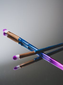 Vibrant makeup brushes with pink and blue handles resting on a reflective surface.