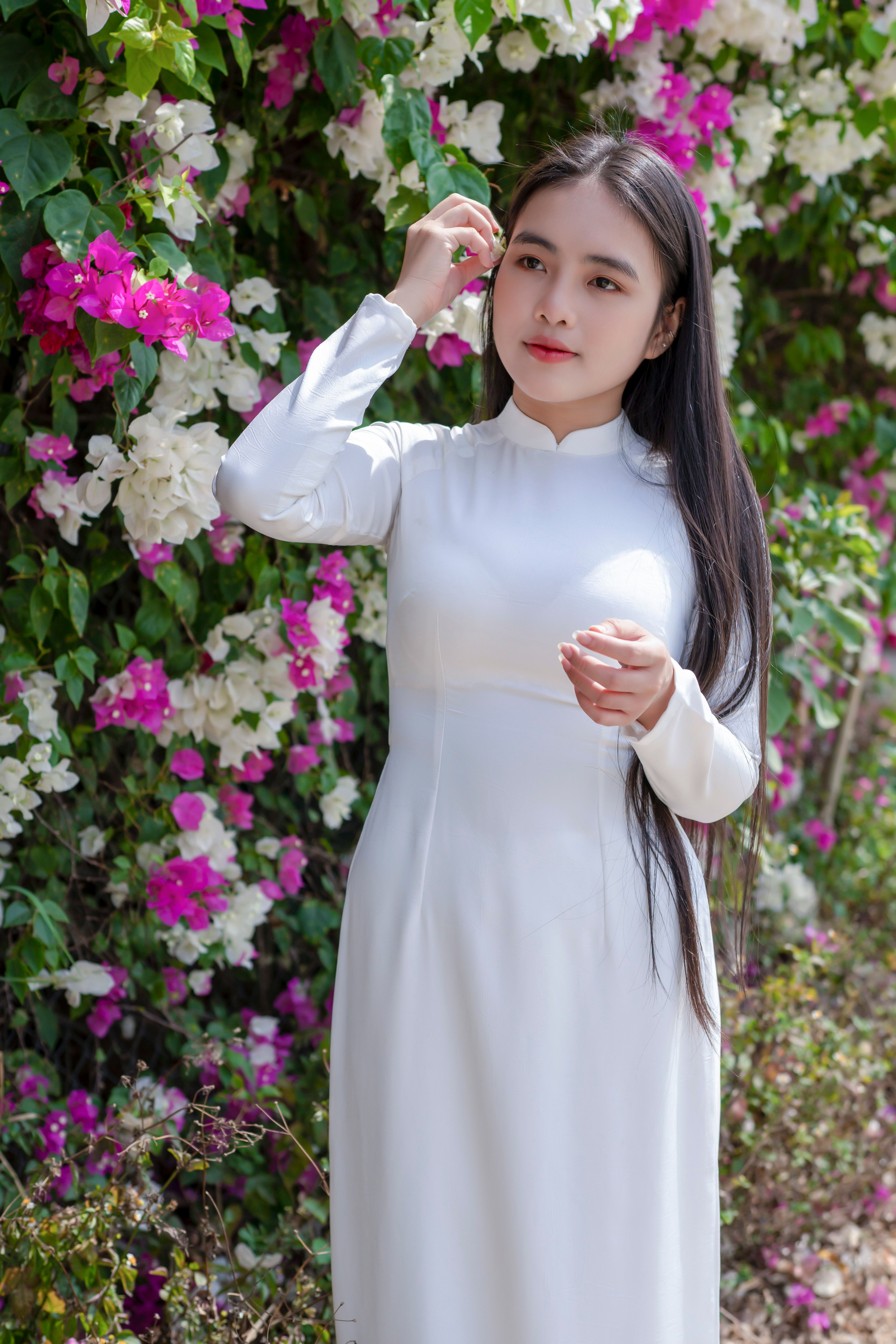 A young woman posing in a white ao dai surrounded by vibrant bougainvillea flowers.