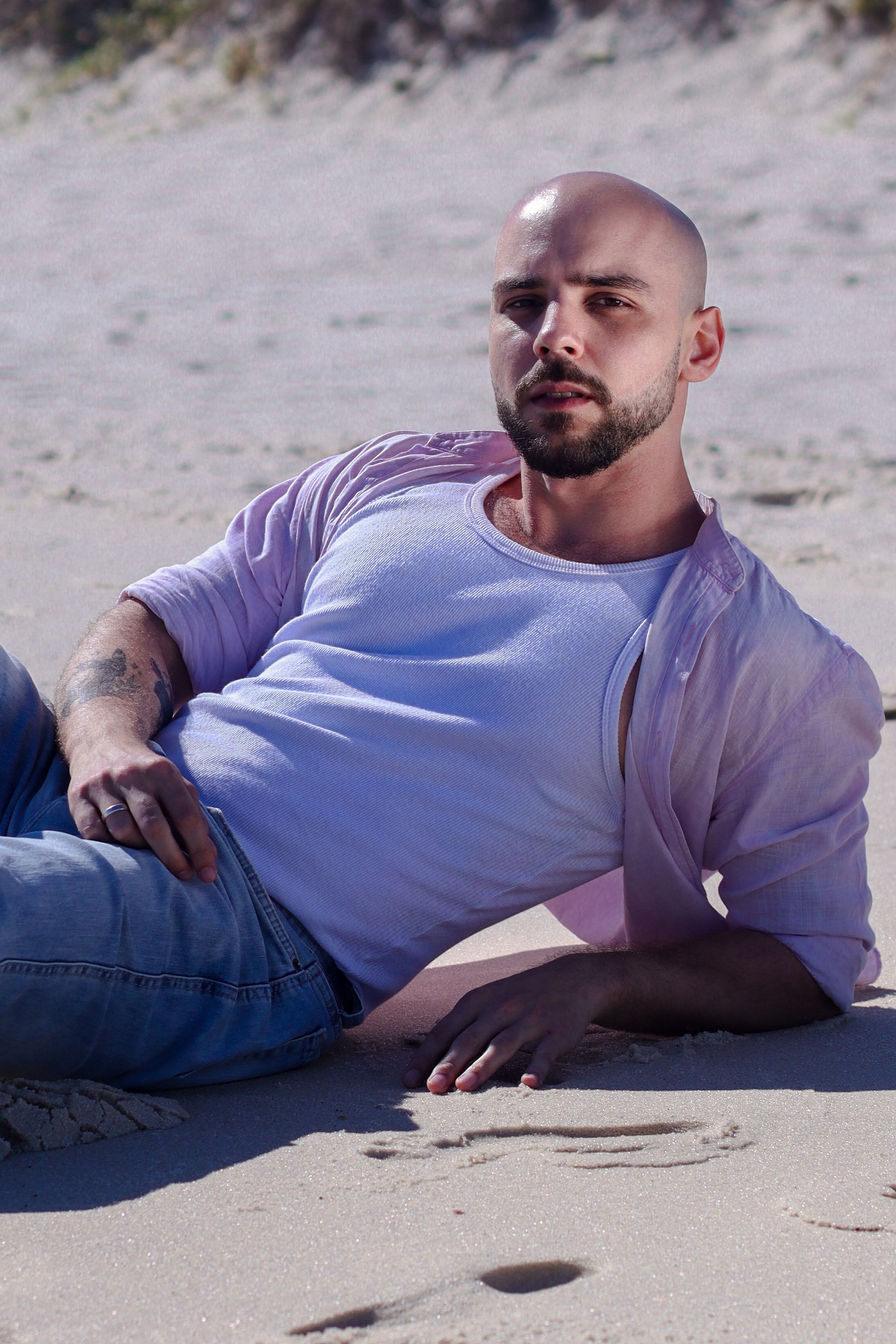 Bald man with a beard relaxing on a sunlit sandy beach, exuding calm and leisure.