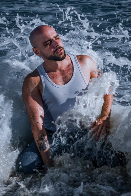 Free A Man in a Tank Top Sitting on the Shore in Splashing Waves  Stock Photo