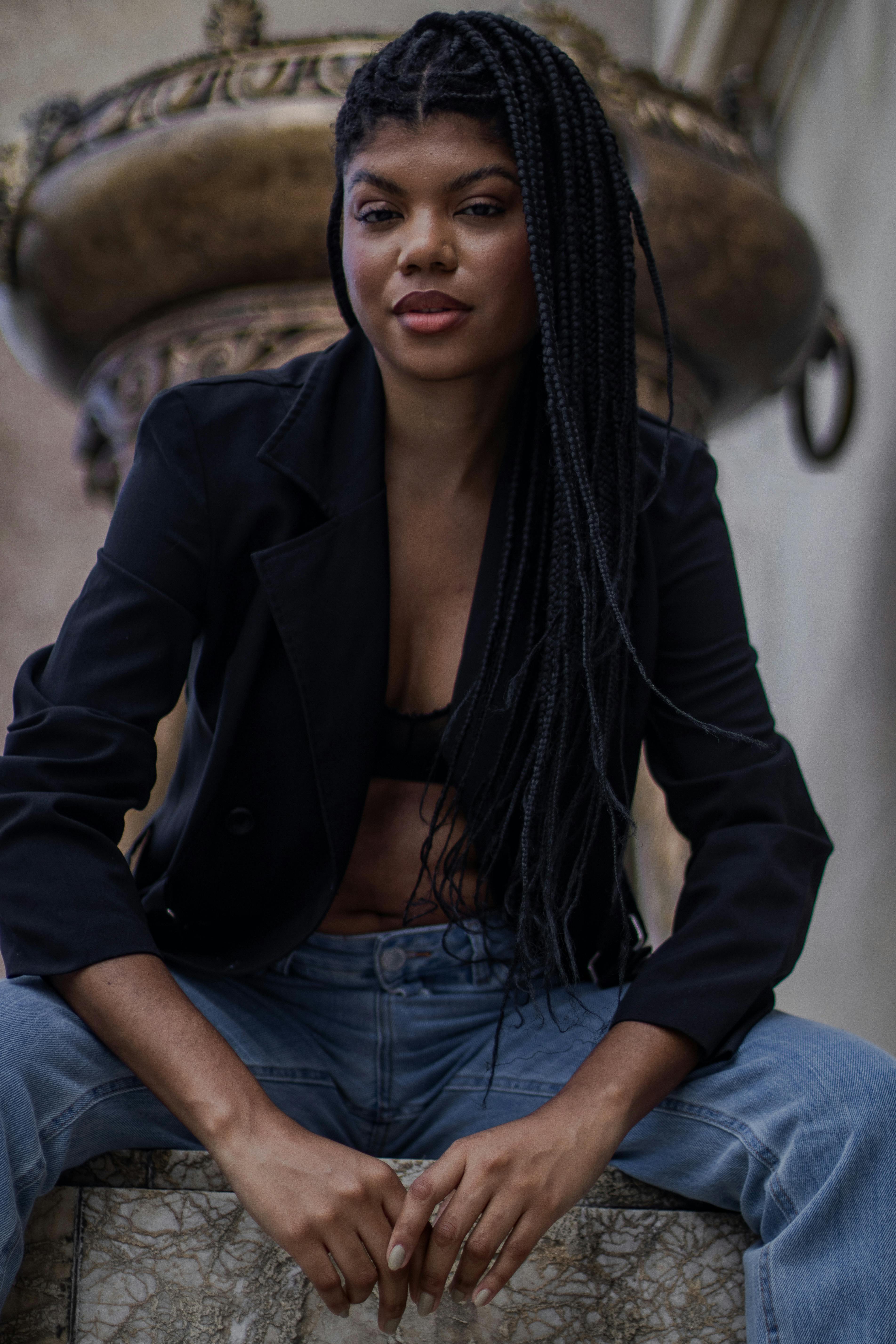 Fashionable young woman with braided hair sitting confidently, wearing a black blazer and jeans.