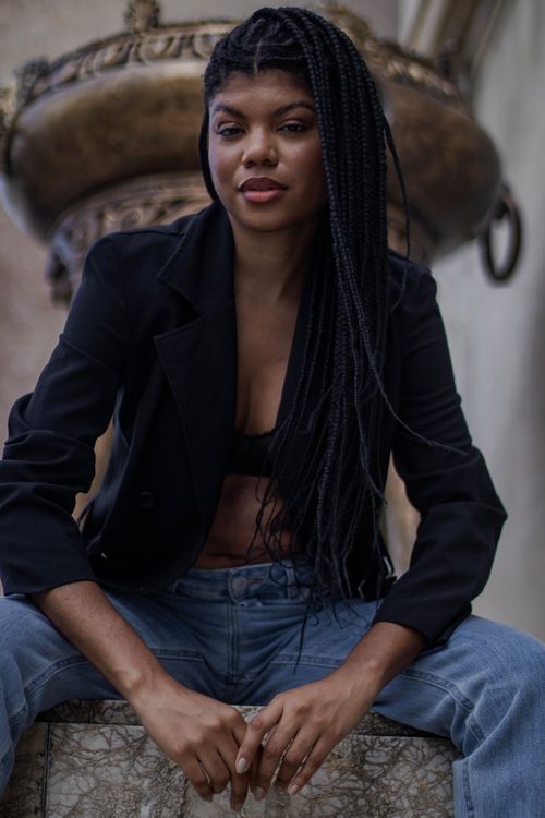 Free Fashionable young woman with braided hair sitting confidently, wearing a black blazer and jeans. Stock Photo