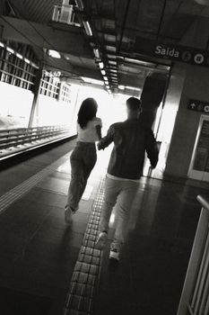 Man and woman running hand-in-hand on a railway platform in black and white.