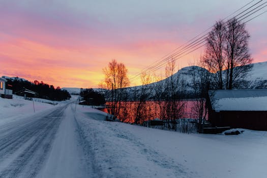 A peaceful winter scene in Laksvatn, Norway, featuring snow-covered roads and a vibrant sunrise over scenic mountains.