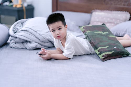 A young boy laying on a bed with a pillow and a remote control