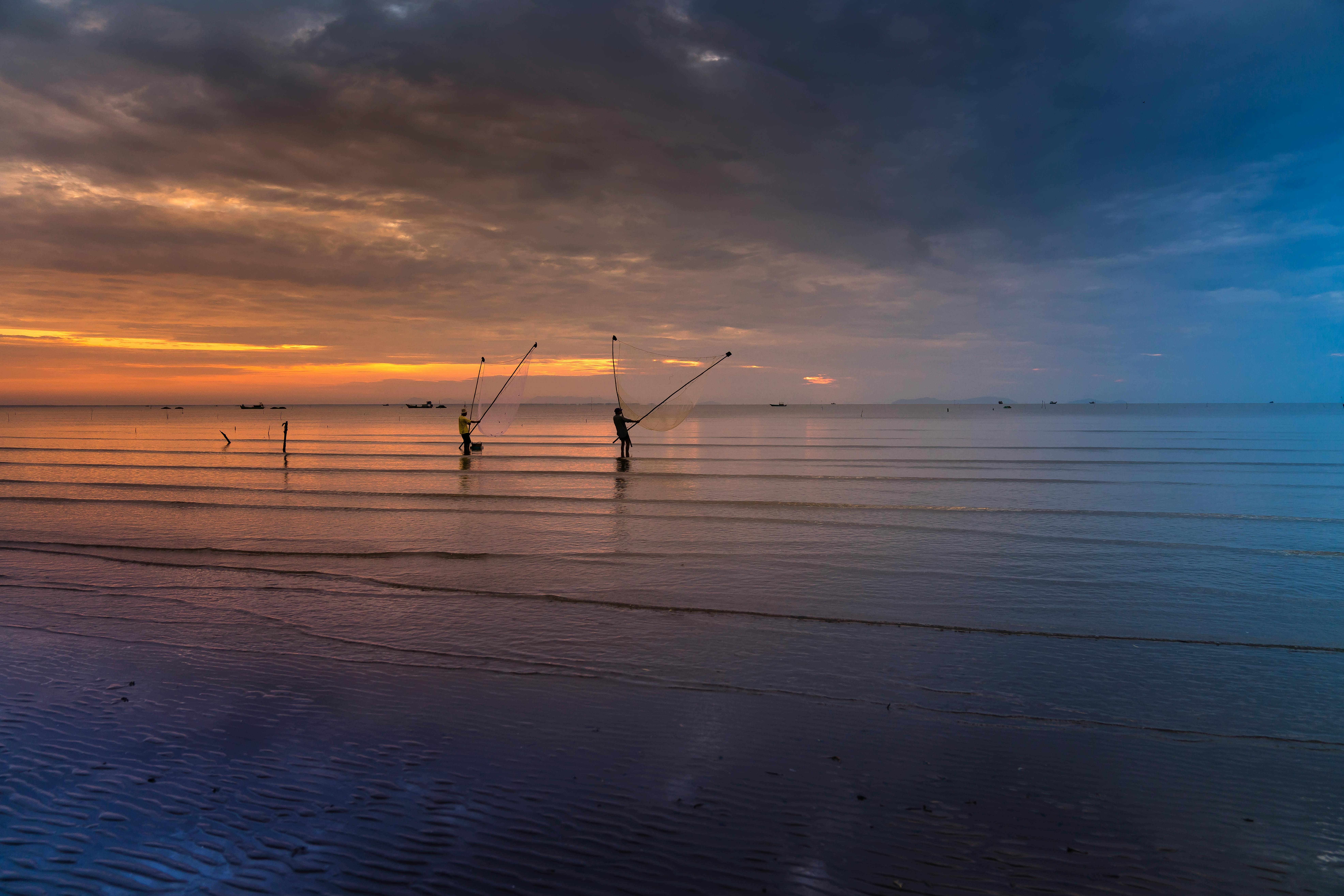 两个渔民在平静的海滩上，迎着充满活力的日落向大海撒网