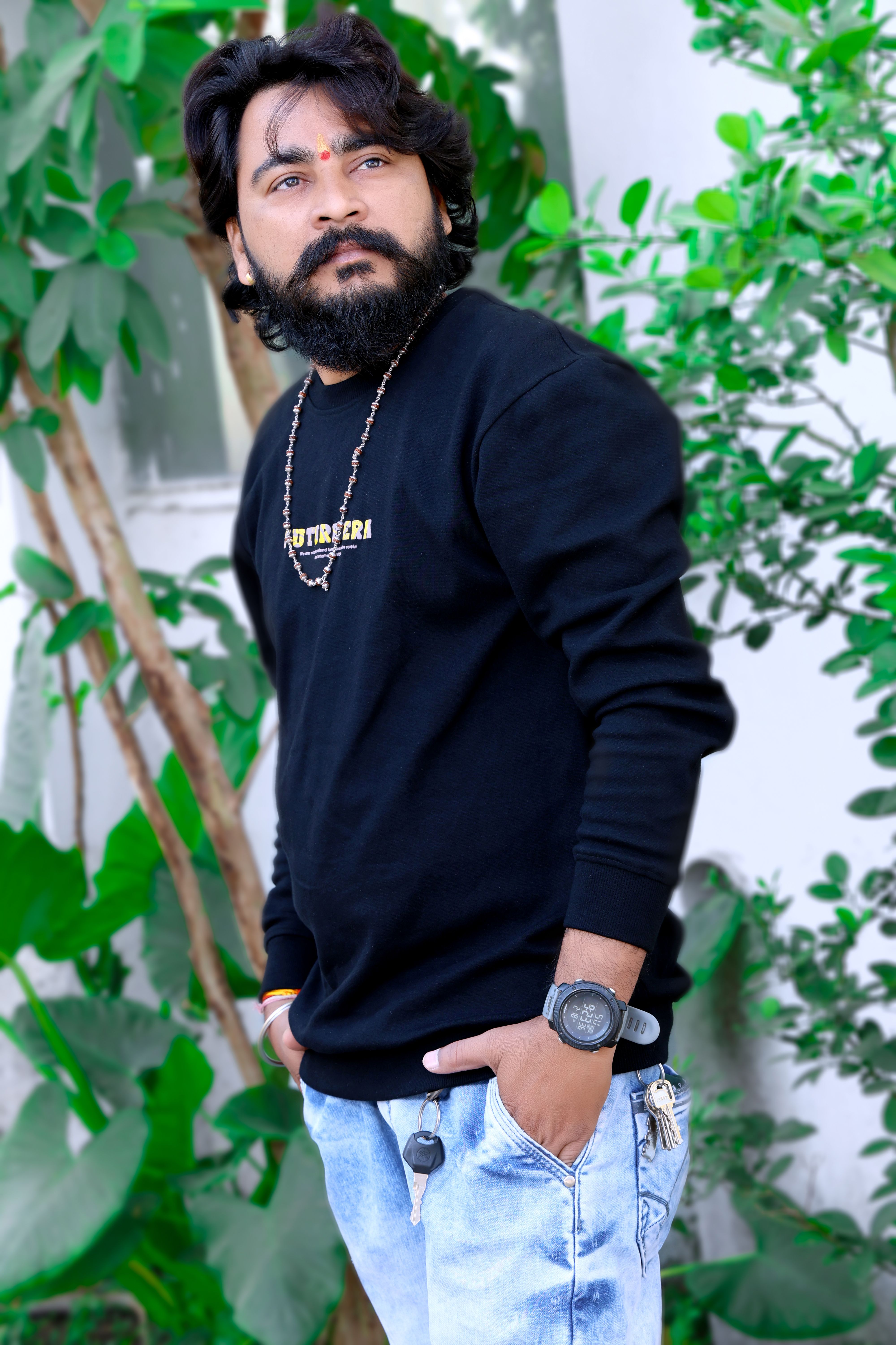 Fashion portrait of a bearded man in a black pullover, outdoor setting with greenery.