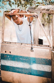 一位年轻男模在阳光明媚的户外，倚着一扇质朴的车门摆出姿势，展现出时尚轻松的风貌