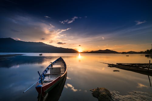 Free Tranquil sunset view with a boat on a calm lake reflecting mountains and golden sky. Stock Photo