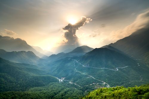 Free Scenic view of mountain range with winding road and sunrays breaking through clouds during sunset. Stock Photo