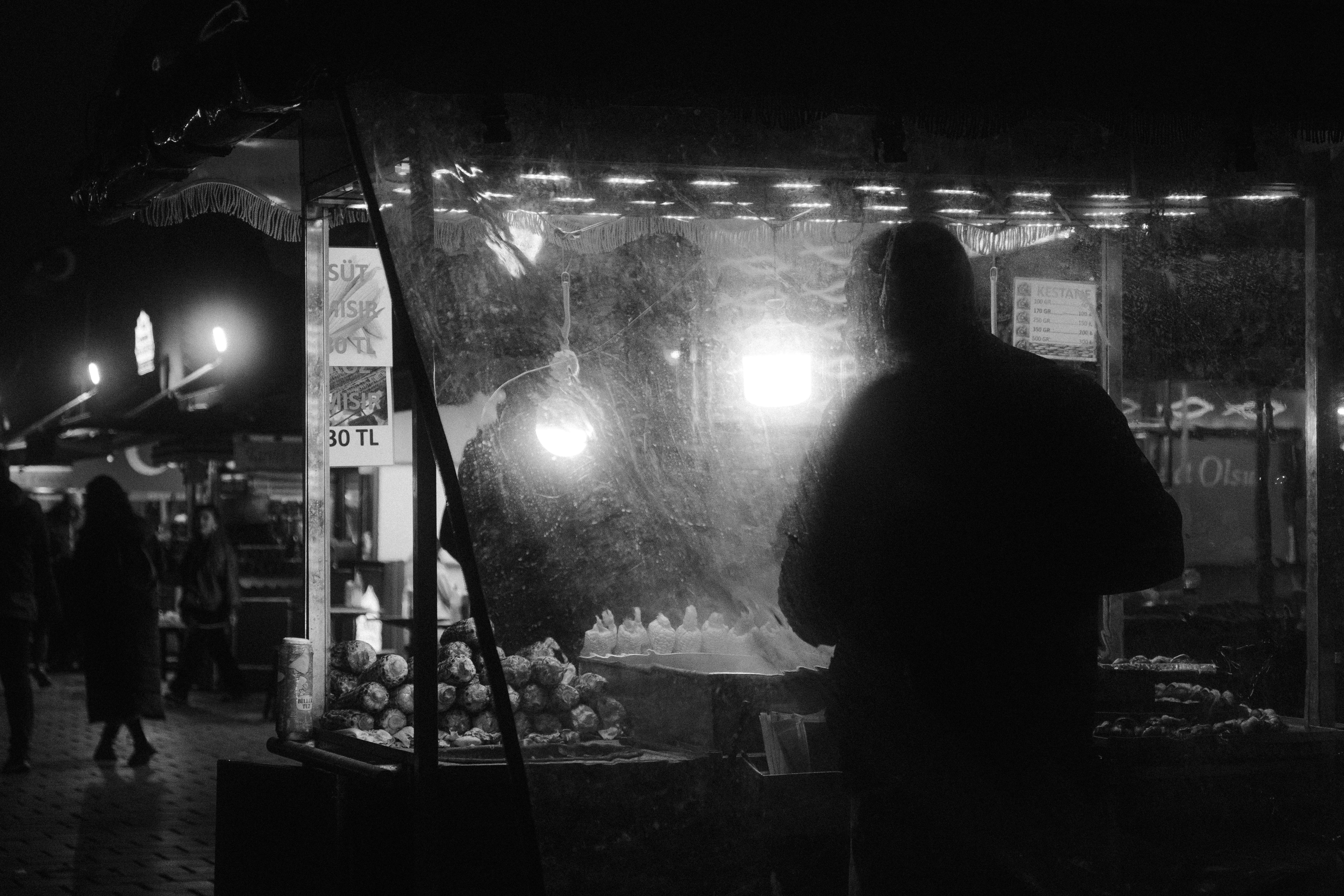 A black and white image of a person silhouetted at a night market stall offering street food.