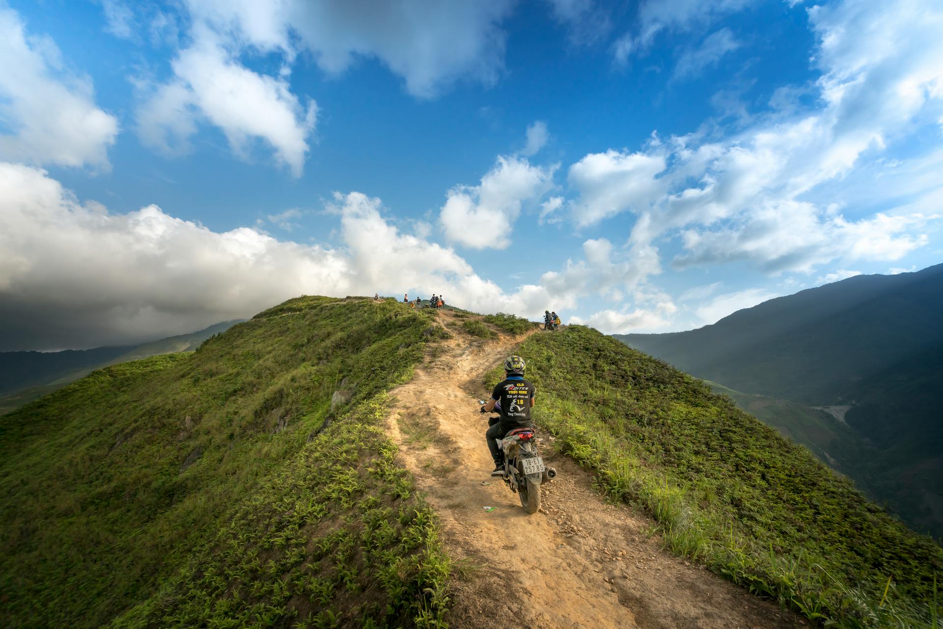 A motorcyclist travels along a rugged mountain path under a vibrant summer sky, perfect for adventure enthusiasts.