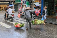 繁华的城市街道上，一个戴着传统帽子的街头小贩正携带着新鲜蔬菜