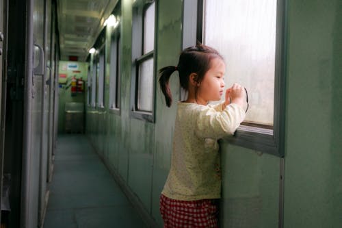 Photo of Girl Standing by Window in Train Hallway Looking Outside