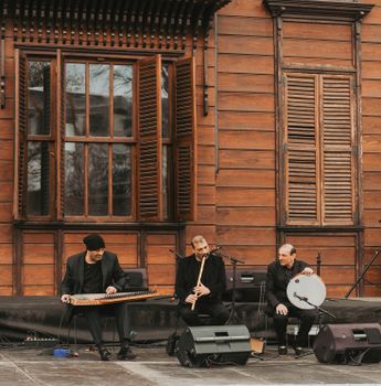 Musicians play traditional instruments in an outdoor setting against a rustic wooden backdrop.