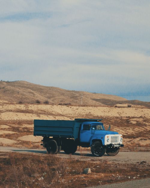 Free A solitary blue truck parked on a rural road with rolling hills in the background, under a cloudy sky. Stock Photo