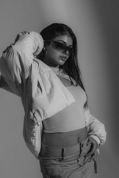 Stylish black and white portrait of a woman in a studio, modeling contemporary fashion with sunglasses.