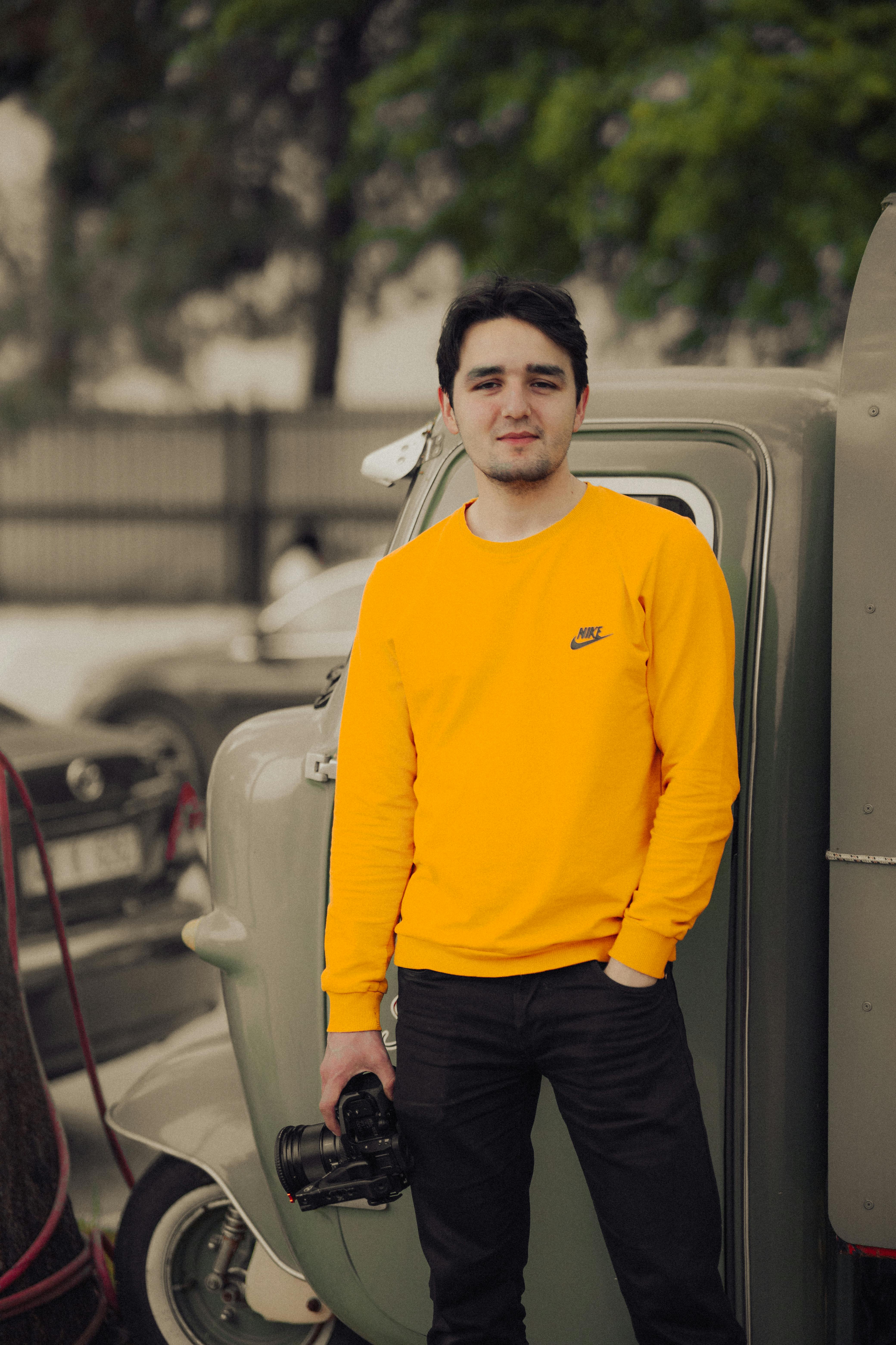 Young man in yellow pullover holding a camera, standing by a vintage car outdoors.