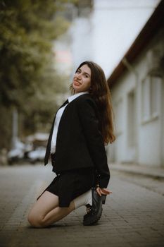 A fashionable woman kneels and smiles on a city sidewalk, wearing a suit jacket and showcasing urban style.