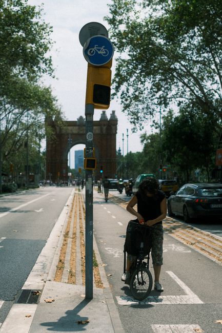 A woman adjusts her bicycle in a bike lane with the Arc de Triomf in the background, Barcelona.