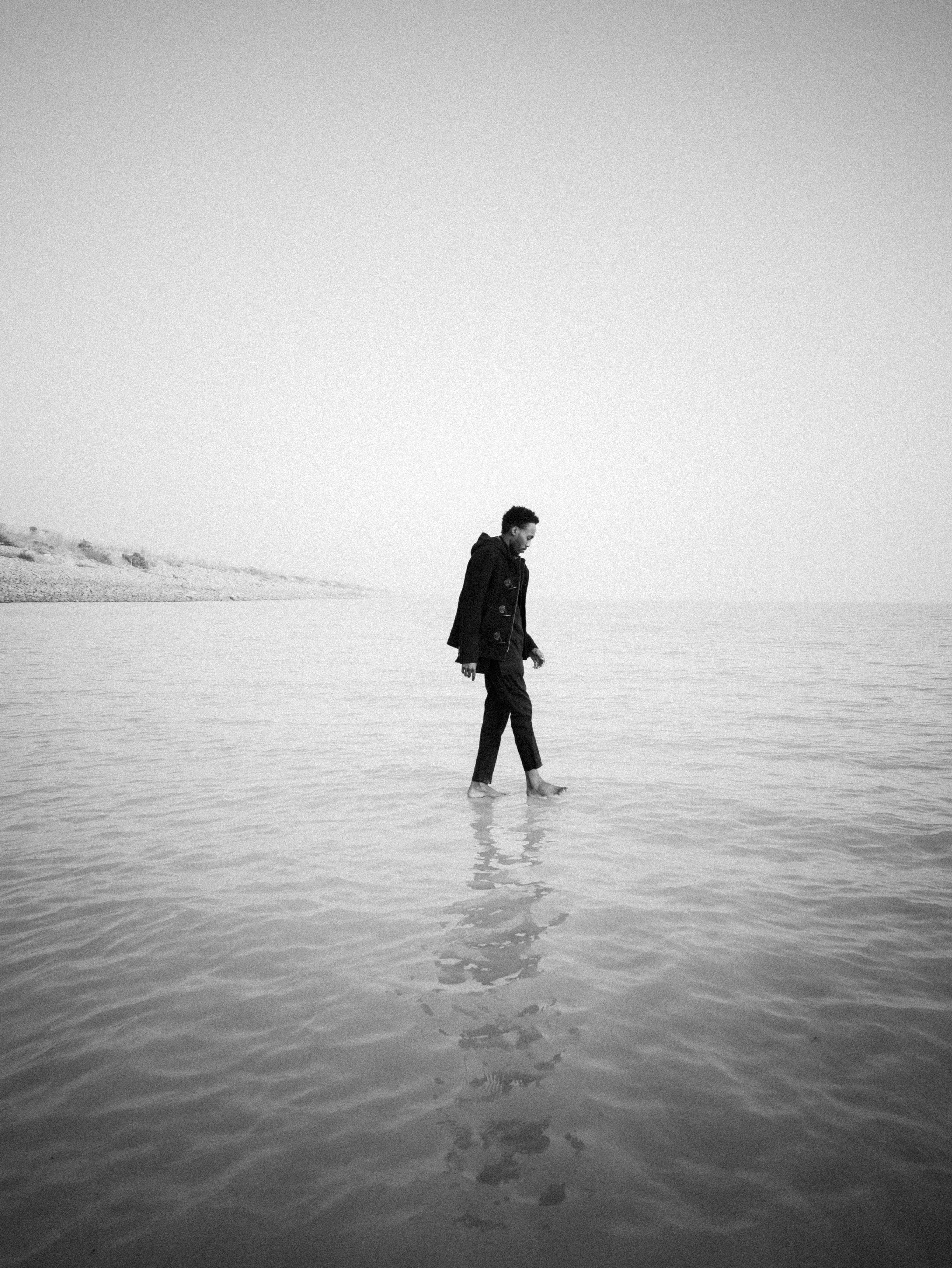 A man walks barefoot along the calm seaside in a peaceful black and white scene.
